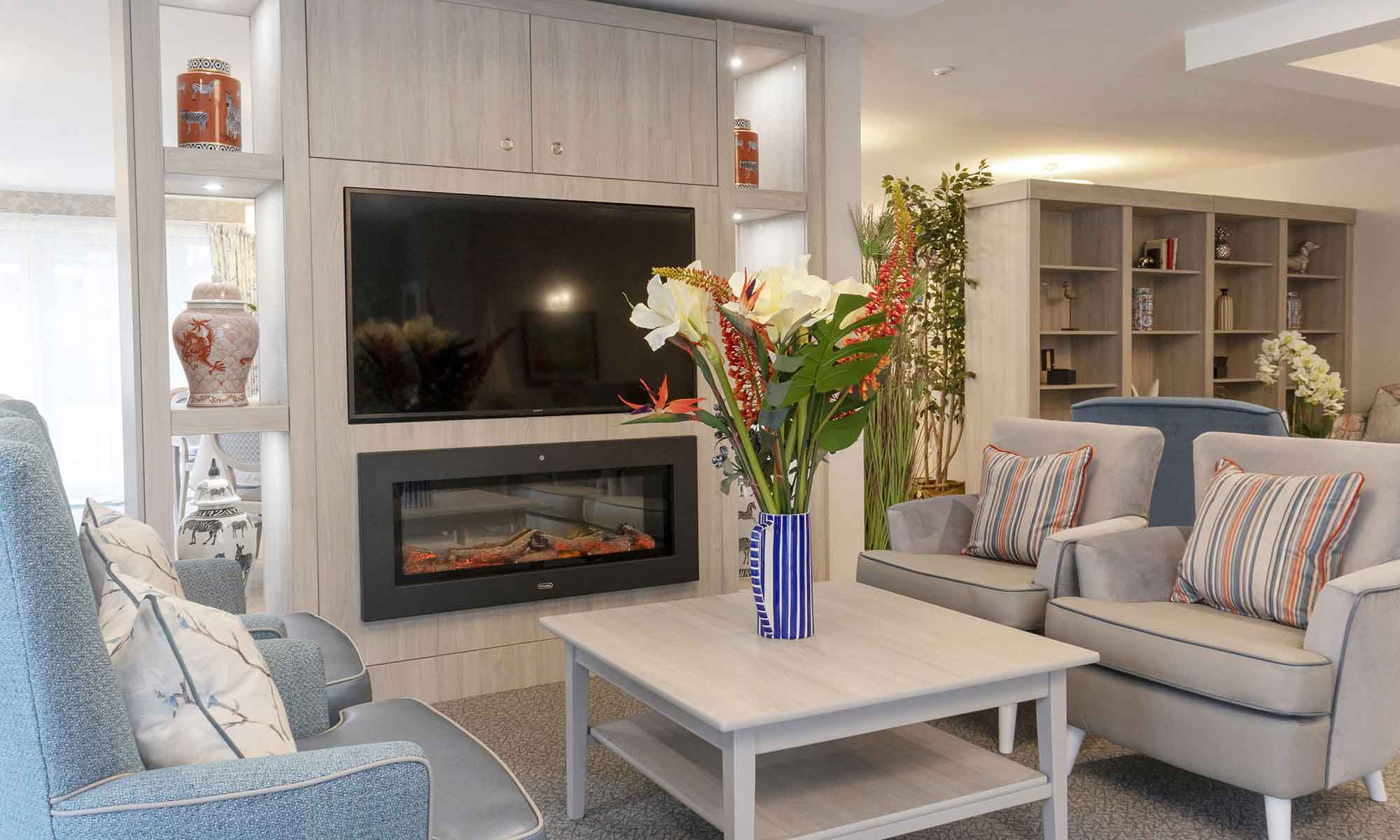 A residents' lounge at a St Mary's care home. Bespoke cabinetry with an inbuilt fire and TV. The Lubeck square coffee table with a flower arrangement in a blue striped jug and 2 Saluzzo high back and 2 Saluzzo low back chairs.