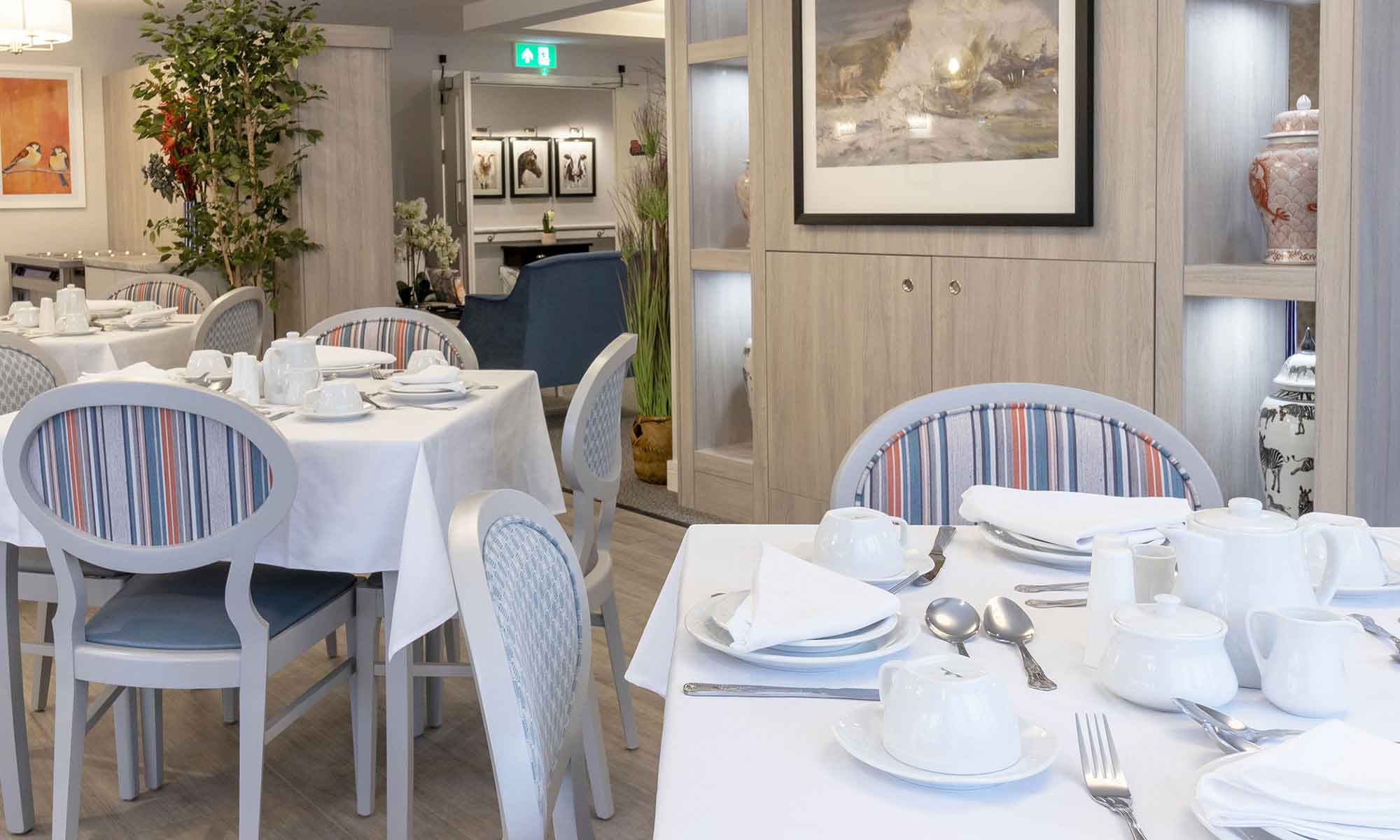 A dining room interior at St Mary's Care Home . A close up of a dining table set up with white tableware and silver cutlery. Featuring a Sesto side chair upholstered in a contrast stripe fabric. A large piece of artwork above bespoke in-built storage cupboards.