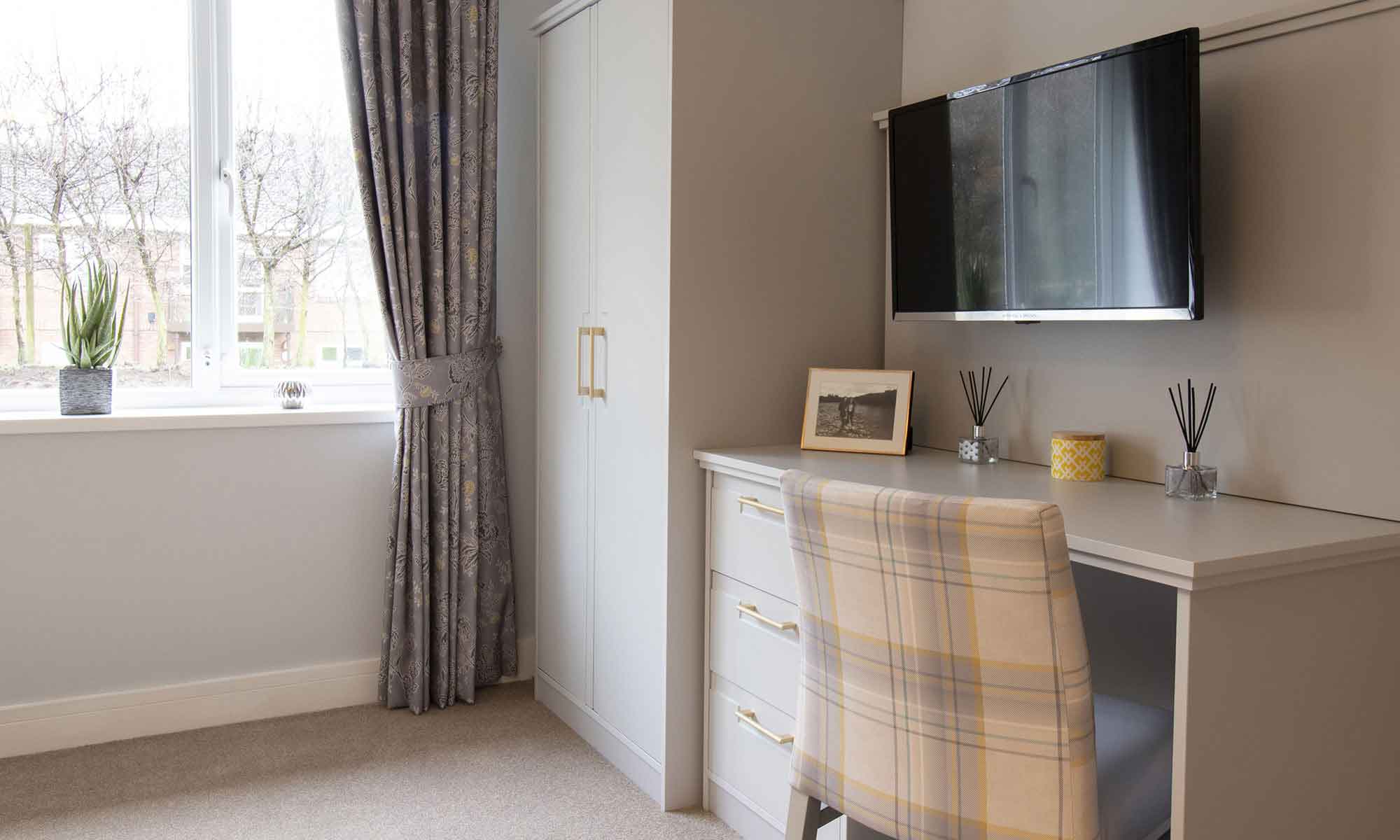 An image of a bedroom interior at St Mary's Care Home in Anlaby. The Windsor double wardrobe and dressing table in light grey with gold metallic handles. A dressing table chair in soft grey and mustard check fabric.