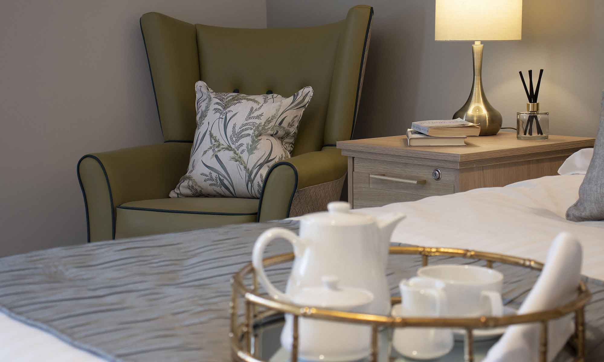An image of a bedroom interior at St Mary's Care Home in Anlaby. A Valencia wingback chair in the corner in a olive green fabric with contrast piping and scatter cushion, the Windsor bedside cabinet with a gold lamp, book and reed diffuser on top with a close up of the bed with a tray with a white tea set on top.