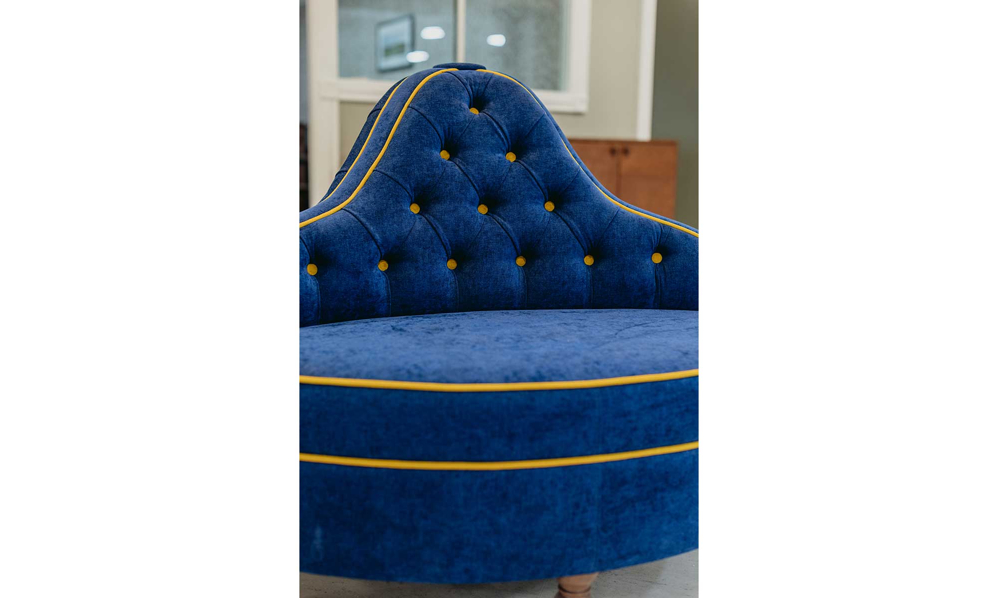 A close up of the ashbourne ottoman in a blue velvet with yellow piping