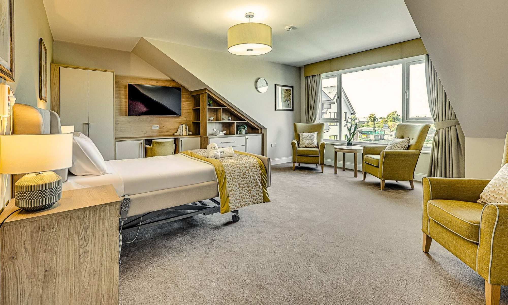 Luxury care home bedroom featuring an adjustable bed with upholstered headboard, integrated fitted cabinetry and shelving, and a seating area with two armchairs near a large window. Soft neutral tones with mustard accents create a warm and calming atmosphere. Furniture designed and manufactured by Shackletons.