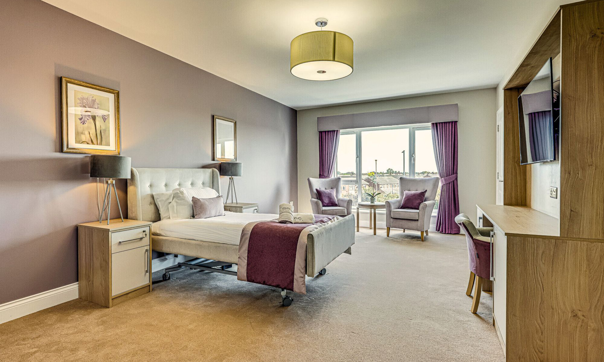 Elegant care home bedroom with upholstered bed, coordinating bedside cabinets, and a seating area overlooking large windows. Interior palette in soft lavender and mauve tones with matching curtains and cushions. Bespoke furniture and cabinetry by Shackletons.