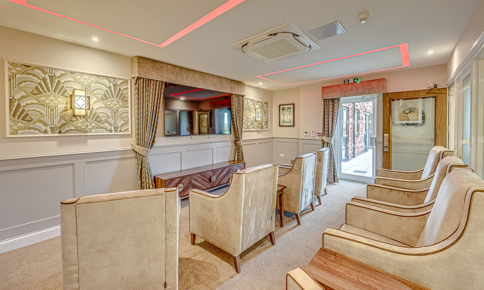 Dedicated cinema room in a care home featuring high-back armchairs, panelled wall detailing, feature wallpaper and a large wall-mounted screen. Warm lighting and soft fabrics create a comfortable, boutique cinema experience. Furniture by Shackletons.