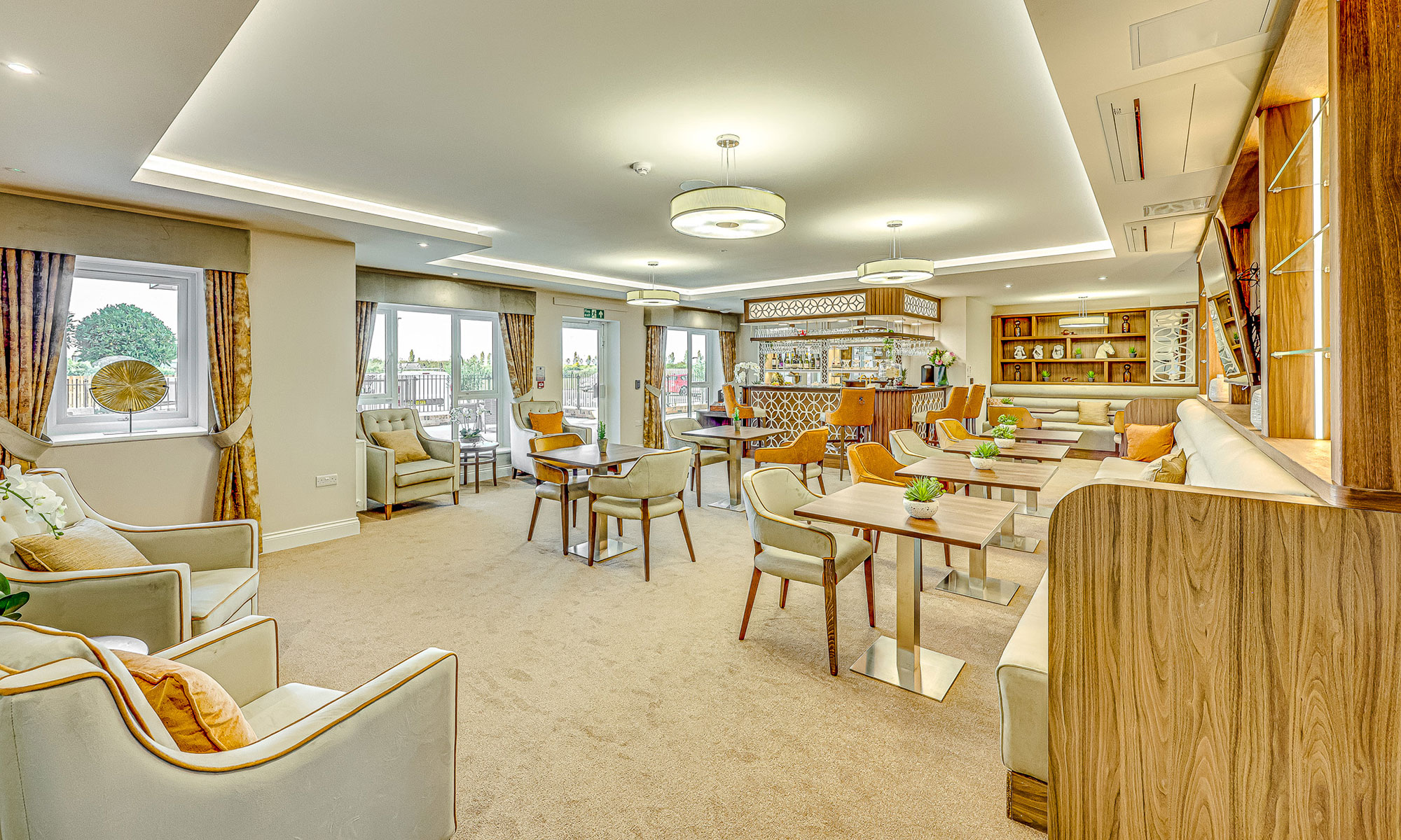 Stylish care home bar and lounge with upholstered dining chairs, sofas, and bespoke joinery. Central bar area with illuminated shelving, wooden accents and comfortable seating zones for socialising. Furniture by Shackletons.