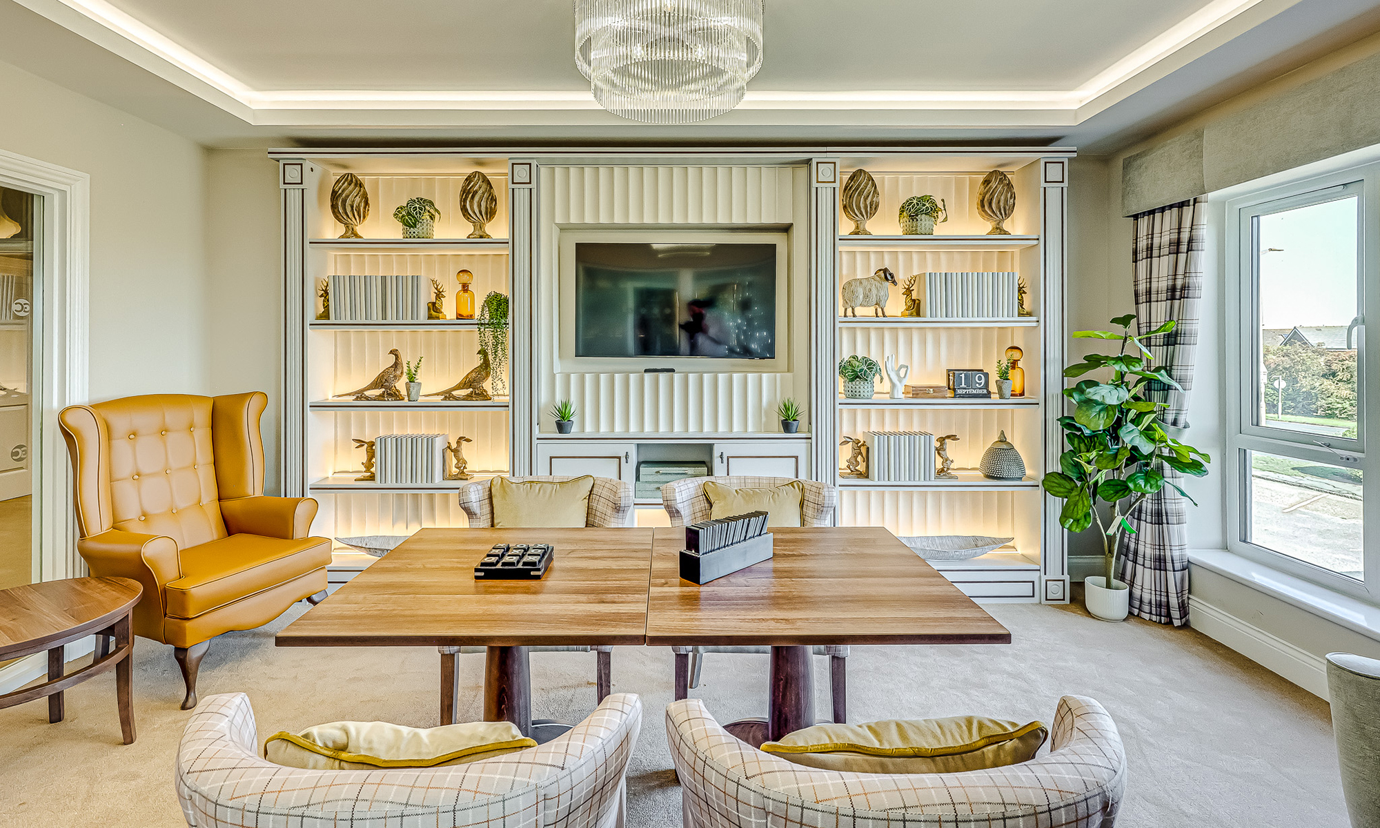 Library-style quiet lounge featuring illuminated built-in cabinetry, upholstered wingback chair, soft seating and wooden games table. Warm neutral colour palette with touches of mustard and plaid fabrics. Furniture by Shackletons.