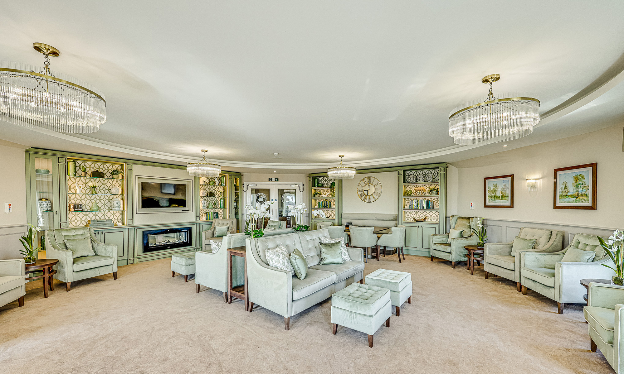 Spacious circular lounge with multiple seating areas, sage green armchairs and sofas, upholstered ottomans and feature shelving with integrated lighting. Elegant and calming space designed for resident relaxation. Furniture by Shackletons.