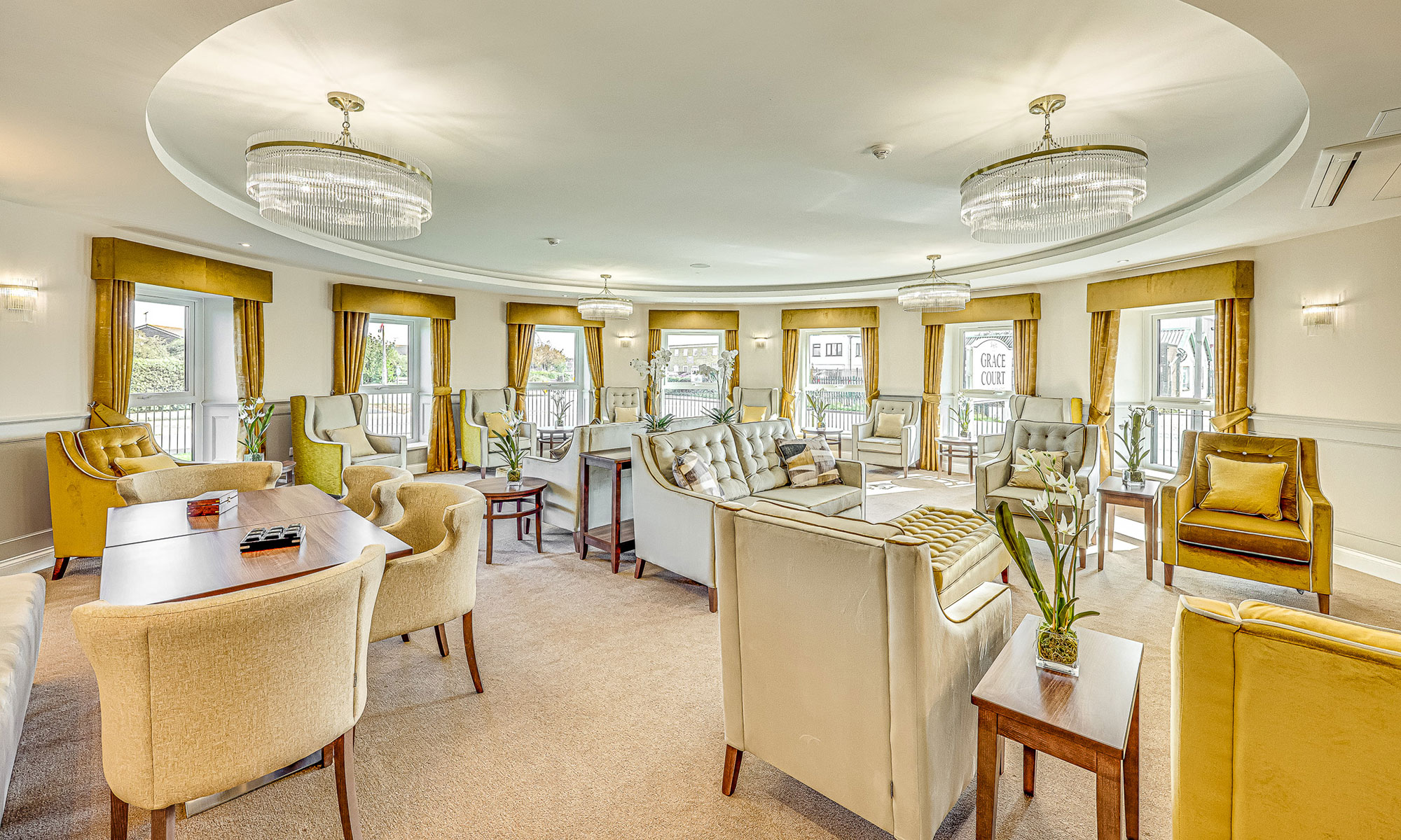 Large circular lounge with gold accent fabrics, high-back chairs, sofas and occasional tables arranged for social interaction. Floor-to-ceiling windows provide natural light. Sophisticated interior completed with Shackletons furniture.