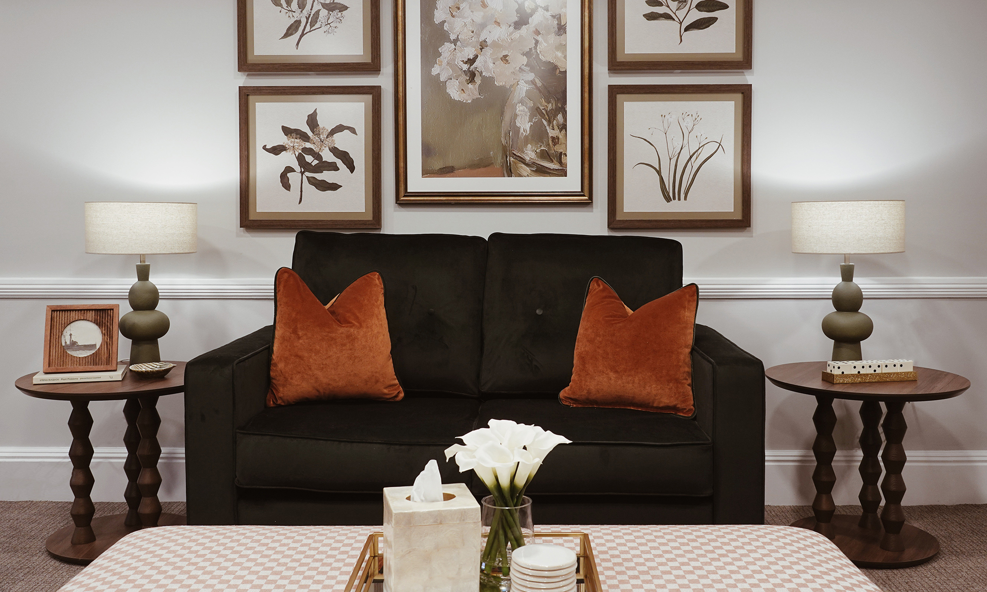 Brompton two-seater sofa upholstered in deep green velvet with rust accent cushions, styled with side tables and lamps at Purley Gardens care home.