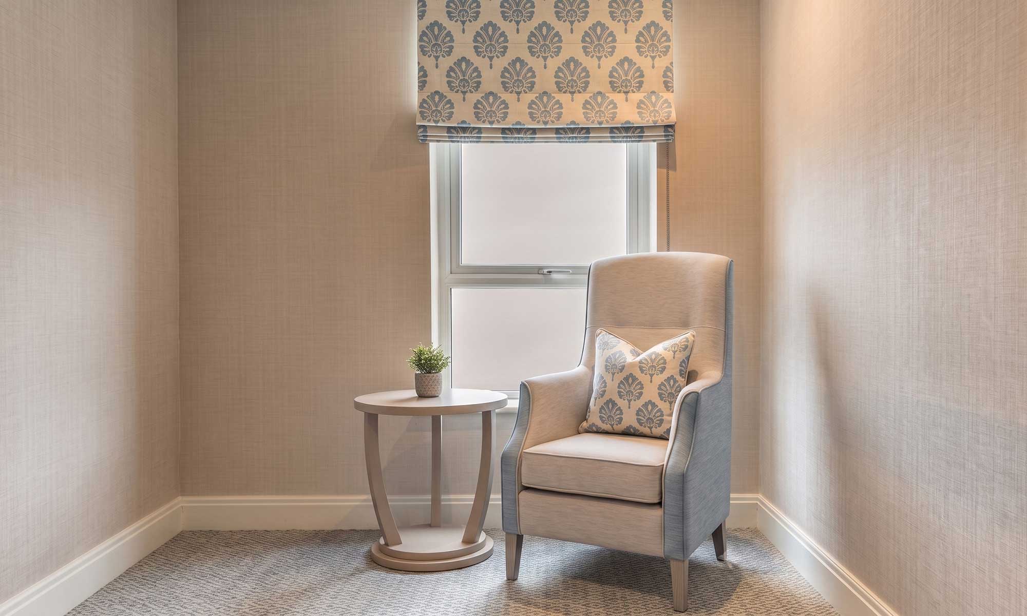 Care home reading area with a patterned Roman blind, high-back chair and matching scatter cushion.
