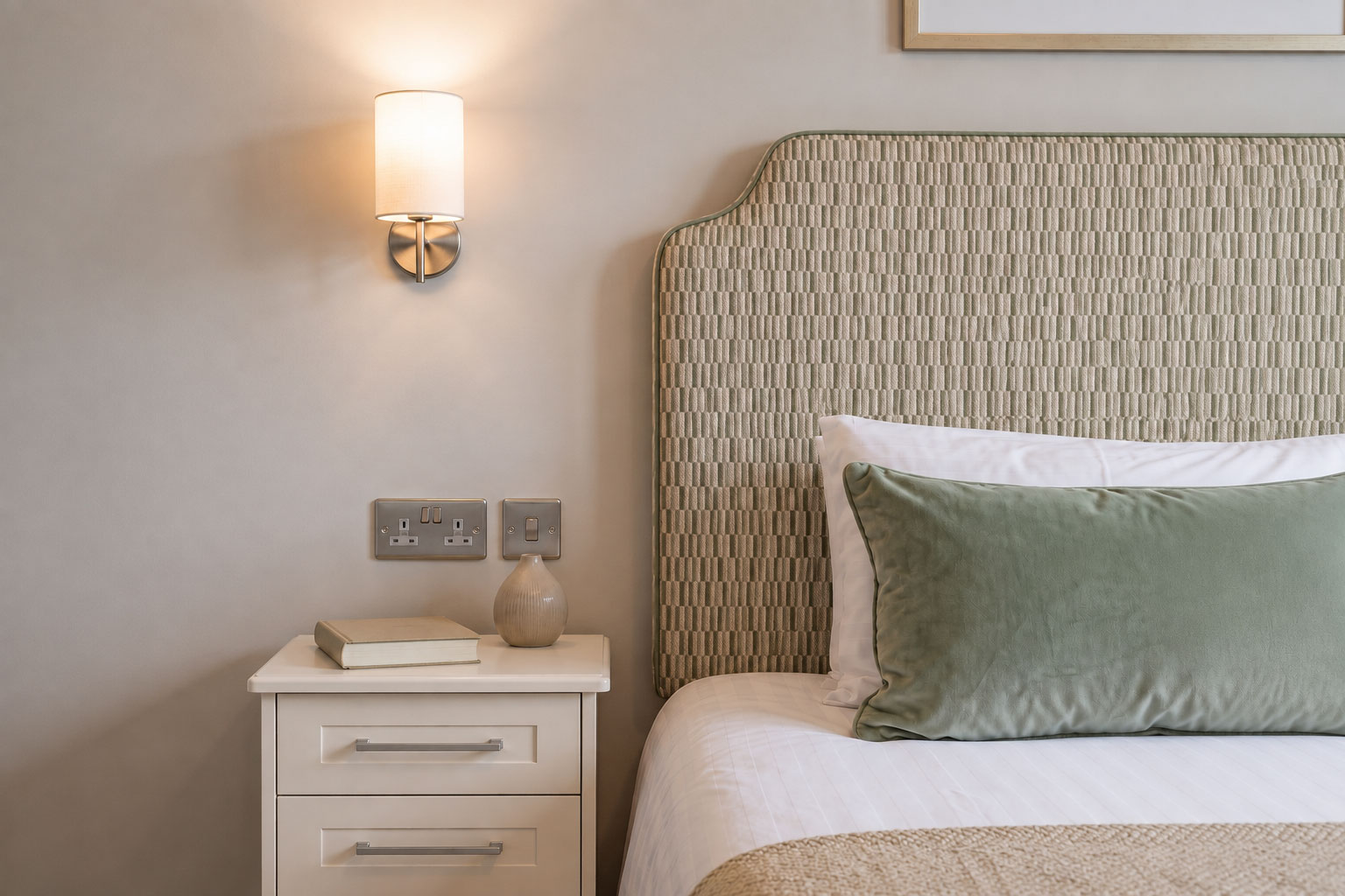 Care home bedroom with a patterned upholstered headboard, sage cushion, white bedside cabinet and wall-mounted reading light.